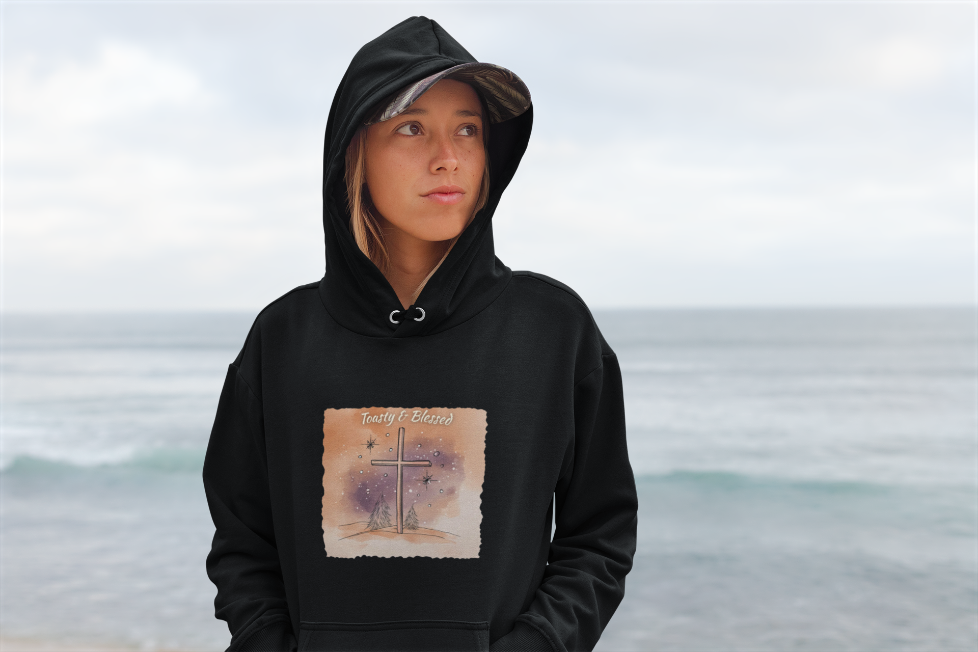Person wearing a black hoodie with a graphic watercolor cross design on a beach